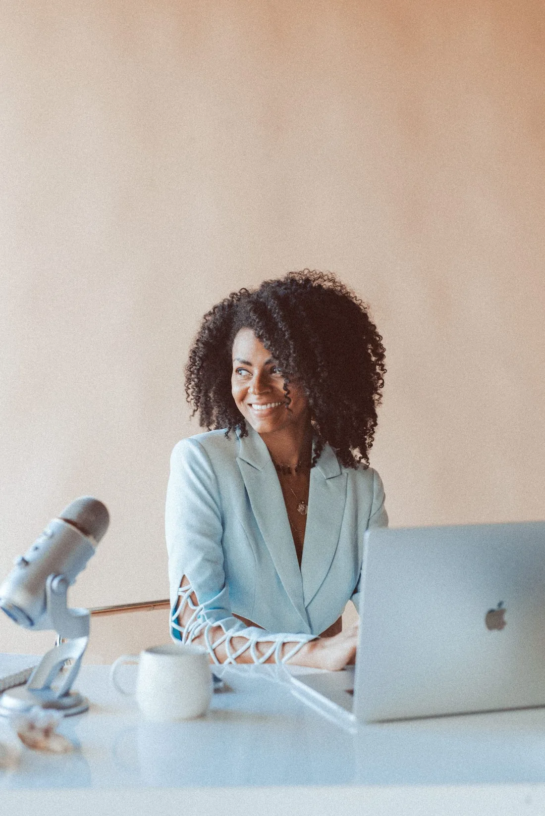 Anna at her desk in a sky-blue blazer, smiling, beside a microphone and laptop.