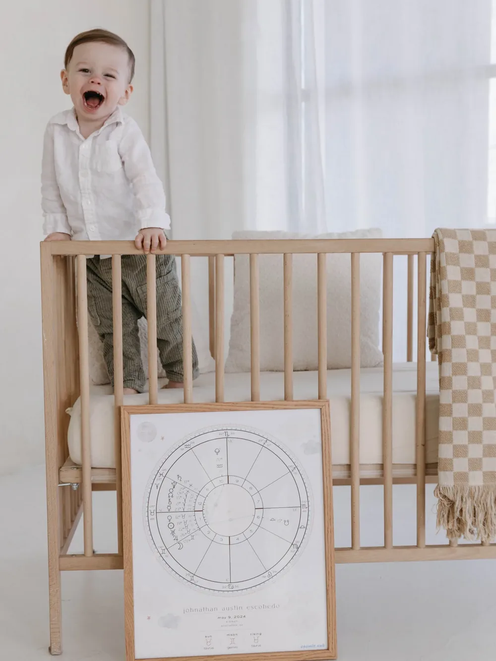 A toddler standing in a crib above a framed baby birth chart print.